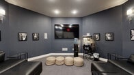 Located at 7999 Wildflower Ln in Columbus, Ohio, this dedicated screening room delivers a true cinematic experience within a cozy, well-appointed single-room layout. The space is finished with deep slate-blue walls throughout, creating a moody, theater-like ambiance that is both intimate and immersive. Recessed ceiling lights provide overhead illumination, while wall-mounted sconce fixtures cast a warm, accent glow along the seating walls — a detail that reinforces the classic movie house atmosphere.

A large flat-screen television is mounted prominently on the main feature wall, flanked by in-wall speaker panels that suggest a capable audio setup. Seating options are varied and flexible, including a sleek black leather sectional sofa complemented by woven pouf ottomans arranged toward the screen. Glass-topped side tables offer a practical surface for refreshments.

A vintage-style popcorn cart and a water cooler add authentic concession-stand character to the room. Framed black-and-white portraits of classic Hollywood actors line the walls, reinforcing the cinematic theme throughout.

Near the entrance, a set of white double doors is paired with a built-in cabinetry station featuring upper cabinets and a dark countertop — a convenient refreshment or storage area. Neutral grey carpeting grounds the space, tying together the dark, polished aesthetic of this purpose-built entertainment room.