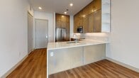 This contemporary single-floor residence offers a thoughtfully designed layout with clean lines and modern finishes throughout. The open-concept living and dining area is anchored by rich hardwood-style flooring and a ceiling fan, while a generous bank of floor-to-ceiling windows floods the space with natural light and provides views of the surrounding urban streetscape. A ceiling fan adds both style and function to this bright, airy living space.

The kitchen is a standout feature, boasting warm taupe shaker-style cabinetry that extends to the ceiling, stainless steel appliances including a double oven range and built-in microwave, and a large peninsula with a sleek quartz countertop and undermount sink. Open shelving adds a contemporary touch and enhances visual openness in the cooking area.

The primary bedroom is generously proportioned, featuring three tall windows with roller shades that invite abundant natural light and urban views. The en-suite bathroom offers a dual-sink vanity with taupe cabinetry and quartz counters, and leads directly to a well-organized walk-in closet. A secondary bedroom features plush neutral carpeting and a large window with cityscape views. Throughout the residence, neutral tones and quality materials create a cohesive, modern ambiance.