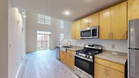 This single-floor residence showcases an open, airy design defined by dramatically high ceilings that lend a sense of generous vertical space throughout. The kitchen is a functional centerpiece, outfitted with light maple cabinetry, granite-style countertops, a farmhouse sink, and a full suite of stainless steel appliances including a gas range, over-the-range microwave, dishwasher, and refrigerator. White subway tile backsplash adds a clean, modern touch, while recessed lighting illuminates the workspace. The kitchen flows seamlessly into the living area, where soaring ceilings and large windows—including transom-style clerestory windows—flood the space with natural light and offer views of the surrounding urban streetscape. A private balcony extends the living area outdoors. Bedroom spaces feature soft carpeting, tall ceilings, and well-sized windows that continue to bring in natural light. Closets are equipped with built-in rods and shelving for practical storage. The bathroom is finished with matching maple cabinetry, granite-topped vanity, and light wood-look flooring. A stacked in-unit washer and dryer adds everyday convenience.
