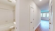 This single-floor unit offers a clean, modern aesthetic defined by consistent hardwood flooring, crisp white walls, and abundant natural light throughout. The kitchen is a functional highlight, featuring white shaker-style cabinetry with brushed nickel hardware, light granite countertops, a stainless steel undermount sink, and a full suite of stainless steel appliances including an electric range, over-the-range microwave, and dishwasher. A window above the counter brings in natural light and frames views of the surrounding cityscape. The living area is bright and open, anchored by a pair of large windows that offer expansive urban views, filling the space with daylight and a strong sense of connection to the city. The bedrooms are well-proportioned, each finished with hardwood floors and featuring closet storage — one with a sliding door closet and built-in rod, another with a reach-in closet. A hallway connects the main living areas to the private spaces, leading to a full bathroom finished with white subway tile in the tub-shower surround, light tile flooring, a white vanity with a clean countertop, and a framed mirror. The overall design is cohesive, neutral, and move-in ready.