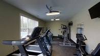 This fitness center offers a practical and well-organized space designed to support a range of workout routines. The room is filled with natural light streaming through large windows fitted with horizontal blinds, creating a bright and energizing environment. The neutral, light-toned walls contribute to an open and clean aesthetic throughout the space.

The cardio section is prominently featured, housing multiple treadmills and elliptical machines, providing ample options for aerobic exercise. Toward the back of the room, a multi-function strength training station offers a variety of resistance-based exercises, complemented by additional free-standing equipment suitable for targeted muscle training. The flooring features a durable, textured surface designed to withstand heavy equipment and high-traffic use.

Overhead, two ceiling fans with integrated lighting fixtures help maintain comfortable air circulation during workouts. The space is efficiently laid out to maximize usability while maintaining clear pathways between equipment. The overall ambiance is functional and inviting, making it a convenient and accessible fitness resource. Whether focused on cardiovascular conditioning or strength building, this fitness center is equipped to meet a wide variety of exercise needs.