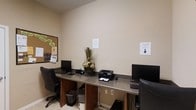 This single-floor business center offers a functional and efficiently organized workspace suited for everyday computing and printing tasks. The room features a continuous granite-topped workstation that lines the walls, providing ample desk space for multiple users simultaneously. Two desktop computer workstations are set up with monitors, keyboards, and mice, while a shared laser printer sits conveniently within reach at the center of the counter. Each workstation is paired with ergonomic rolling chairs, ensuring comfort during extended use. A cork bulletin board mounted on the wall serves as an information hub, displaying notices and announcements with colorful labels for easy navigation. A decorative floral arrangement adds a welcoming touch to the otherwise utilitarian space. The walls are finished in a neutral beige tone, contributing to a clean and professional atmosphere. Printed signage is posted on the walls to guide users. Light-toned flooring and adequate overhead lighting keep the space bright and practical. This business center is well-suited for tasks such as printing documents, browsing, or handling correspondence, making it a convenient amenity for guests or tenants seeking a dedicated workspace.