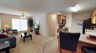 This single-story residence is designed around an open-concept floor plan that seamlessly integrates the living, dining, and kitchen areas into one cohesive space. The living room is carpeted and features a ceiling fan with a light fixture, a large window that allows natural light to brighten the room, and ample space for comfortable seating and entertainment. A flat-screen television is positioned against the wall, complemented by decorative greenery that adds a natural touch to the room.

Adjacent to the living area, the dining space transitions into the kitchen via tile flooring, providing a clear visual distinction between zones. The kitchen is equipped with warm-toned wood cabinetry, black appliances including a microwave, stove, and additional countertop appliances, offering a practical cooking environment. The partial wall separating the kitchen from the main living area adds architectural definition while maintaining the open feel of the layout.

The overall color palette consists of neutral beige and cream tones throughout, contributing to a warm and inviting ambiance. The single-floor layout ensures easy accessibility and efficient use of space throughout the home.