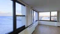 This single-floor space is defined by its striking abundance of natural light and panoramic views. Large, floor-to-ceiling windows wrap around multiple walls, framing broad elevated vistas of the surrounding urban landscape and open sky — a standout architectural feature that immediately draws the eye. The windows are fitted with roller shades, offering flexible light and privacy control throughout the day.

The interior maintains a clean, minimalist aesthetic with crisp white walls and a smooth white ceiling that amplify the brightness flooding in from outside. Warm-toned hardwood flooring runs throughout, adding a natural contrast to the otherwise cool, modern palette. Beneath the windows, a built-in baseboard heating unit runs along the perimeter, ensuring comfort across all seasons while keeping the design streamlined and unobtrusive.

The open layout and corner window configuration create a sense of spaciousness and airiness rarely found in urban settings. The dark-framed windows provide a subtle industrial accent that complements the contemporary finishes. Whether used as a living space, office, or creative studio, this space offers an inspiring, light-filled environment with a strong connection to the outdoors through its expansive glazing and elevated perspective.