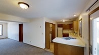 This single-floor residence offers a practical and comfortable layout designed for everyday living. The open-concept design seamlessly connects the kitchen and living area, creating a cohesive and functional space. The kitchen is well-equipped, featuring warm medium-toned wood cabinetry both above and below the counters, a white refrigerator, and a range, providing ample storage and workspace. A peninsula with a double-basin sink and a light-colored countertop extends into the living space, serving as both a prep area and an informal divider between the kitchen and the adjoining carpeted living area. The living area benefits from the open sightlines created by this layout, giving the space an airy, connected feel. A doorway off the main living space leads to a dedicated laundry area, where a washer and dryer are conveniently housed. Flush-mount ceiling light fixtures illuminate the interior throughout. Neutral-toned walls and light carpet in the living area contribute to a clean, understated aesthetic, while warm wood accents in the cabinetry and trim add a touch of coziness to the overall atmosphere.