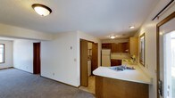This single-floor residence offers a practical and comfortable layout designed for everyday living. The open-concept design seamlessly connects the kitchen and living area, creating a cohesive and functional space. The kitchen is well-equipped, featuring warm medium-toned wood cabinetry both above and below the counters, a white refrigerator, and a range, providing ample storage and workspace. A peninsula with a double-basin sink and a light-colored countertop extends into the living space, serving as both a prep area and an informal divider between the kitchen and the adjoining carpeted living area. The living area benefits from the open sightlines created by this layout, giving the space an airy, connected feel. A doorway off the main living space leads to a dedicated laundry area, where a washer and dryer are conveniently housed. Flush-mount ceiling light fixtures illuminate the interior throughout. Neutral-toned walls and light carpet in the living area contribute to a clean, understated aesthetic, while warm wood accents in the cabinetry and trim add a touch of coziness to the overall atmosphere.