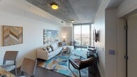 This contemporary high-rise unit offers a thoughtfully designed open-concept layout spread across a single floor. The living area immediately captures attention with its exposed concrete ceiling, a distinctive architectural detail that lends an industrial-chic character to the space. A large sectional sofa anchors the room beneath a striking city-lights canvas, while a vibrant geometric area rug adds a pop of color. Floor-to-ceiling windows flood the living room with natural light and frame expansive panoramic views of the surrounding cityscape and horizon.

The kitchen is outfitted with dark espresso cabinetry, granite countertops, a classic white subway tile backsplash, and a full suite of stainless steel appliances, including a side-by-side refrigerator, electric range, and built-in microwave. A breakfast bar provides additional seating and connects the kitchen seamlessly to the living area.

The bedroom continues the floor-to-ceiling window theme, offering breathtaking elevated views that make for a serene and inspiring retreat. Neutral carpet and warm accent lighting complete the relaxing atmosphere.

The bathroom features dark wood vanity cabinetry topped with granite, a full-width mirror with modern sconce lighting, tile flooring, and a glass-enclosed walk-in shower with white tile surround — a clean and functional finishing touch to this stylish urban residence.