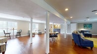 This single-floor clubhouse offers a welcoming and versatile gathering space designed for both relaxed socializing and organized events. The open-concept layout is anchored by rich hardwood flooring that flows seamlessly throughout, tying together the various functional zones with warmth and continuity.

The main lounge area features comfortable seating arrangements, including a neutral sofa and accent chairs accented by bold blue and patterned throw pillows, set atop vibrant chartreuse area rugs. Structural columns frame the space while preserving the open feel, and recessed lighting provides a well-lit, inviting atmosphere.

A dedicated dining area accommodates a round table with contemporary chrome-accented chairs, complemented by wall-mounted artwork and a flat-screen television. Natural light filters in through slatted window blinds, brightening the neutral gray walls.

The kitchenette is a standout feature, showcasing rich dark cabinetry, light stone countertops, and a striking linear mosaic tile backsplash. Stainless steel appliances — including a refrigerator, dishwasher, and beverage cooler — along with pendant lighting over the serving counter make this area well-suited for catering and entertaining. Crown molding throughout adds a refined architectural detail that elevates the overall ambiance of this functional community space.