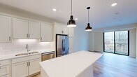 This thoughtfully designed one-bedroom, one-bathroom unit offers a well-appointed layout across a single floor, making efficient use of every square foot. The open-concept kitchen is a standout feature, showcasing crisp white shaker cabinetry with matte black hardware, bright quartz countertops, a classic white subway tile backsplash, and a full suite of stainless steel appliances including a French door refrigerator, electric range, and built-in microwave. A generously sized kitchen island with pendant lighting overhead provides additional prep space and anchors the transition into the living area. Light hardwood flooring runs throughout the main living spaces, complementing the clean, neutral color palette. The living room is bright and airy, highlighted by a large sliding glass door that opens to a private balcony with views of surrounding greenery. The bedroom is comfortably sized, finished with plush carpet flooring and served by a dedicated closet. The full bathroom features a single-sink vanity with quartz countertop, a large mirror, light wood flooring, and a walk-in shower clad in elegant marble-style tile with a sleek black-framed glass enclosure. Recessed lighting throughout enhances the modern, move-in-ready aesthetic of this well-finished unit.