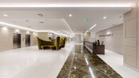 Located at 620 NW Tenth Ave in Miami, FL, this single-floor commercial space presents a refined and professionally designed environment suited for a modern business setting. The expansive lobby serves as the centerpiece, featuring a striking reception desk finished in dark veined marble beneath the "Reflections" signage, set against padded wall panels in warm taupe tones. Polished marble flooring leads visitors through the space, complemented by recessed and linear lighting that creates a bright, welcoming atmosphere.

The layout extends into multiple functional areas, connected by wide, light-filled corridors with terrazzo flooring. Gold-toned accent columns display large-format cityscape artwork, while sculptural metallic ceiling fixtures add visual drama. Glass-enclosed private offices provide transparency while maintaining separation, furnished with workstations and colorful accent seating. A dedicated mailroom area is accessible behind glass partitions.

A standout feature is the vibrant children's play area, decorated with a bold geometric mural, bright green walls, a small slide, bean bag chairs, and a large-screen television. An elevator lobby with padded wall paneling and stainless steel doors offers seamless vertical access. Throughout, the space balances professional functionality with thoughtful, artistic design elements.