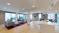 This single-floor space showcases a thoughtfully designed open-plan layout that seamlessly integrates multiple functional zones. The living area is anchored by a plush gray sofa set arranged atop a deep purple area rug, creating a defined lounge space with a comfortable, contemporary feel. A central white structural column punctuates the open floor plan without compromising the sense of spaciousness.

Large floor-to-ceiling windows along the perimeter flood the interior with natural light while offering views of the surrounding urban environment. The flooring throughout is light-toned hardwood, contributing to a clean and airy aesthetic that ties the zones together cohesively.

The kitchen area features a sleek white countertop island with black bar stools, offering a casual seating option that bridges the kitchen and living spaces. Modern pendant lighting accents the kitchen zone. A dining area with additional seating is visible in the background, positioned to take advantage of the natural light from nearby windows.

Recessed ceiling lighting provides uniform illumination throughout, complementing the natural light. The overall ambiance is modern and minimalist, characterized by neutral tones, clean lines, and thoughtful spatial organization.