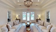 Located at 18172 Section St in Fairhope, Alabama, this thoughtfully designed single-floor space blends coastal elegance with warm, inviting functionality. The grand main lounge serves as the heart of the space, featuring a striking tray ceiling adorned with a rustic orb chandelier, wide-plank hardwood-style flooring, and oversized French doors that flood the room with natural light. Symmetrically arranged neutral sofas, accent tables, and curated wall art create a refined yet comfortable atmosphere. An adjacent sitting room offers a more intimate setting, with four striped accent chairs arranged around a central table and a wall-mounted television, ideal for small gatherings. The expansive kitchen is a standout, showcasing a large shiplap island with seating, crisp white cabinetry, stainless steel appliances, and a beverage station, all illuminated by bold brass pendant lights. The open layout connects seamlessly to a bright dining area, where a round table is bathed in natural light from large windows overlooking a lush green landscape. Crown molding, neutral tones, and consistent hardwood-style flooring unify the space throughout, creating a cohesive and polished environment that is both functional and welcoming.