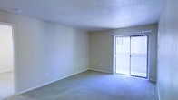 This single-story space features a generously sized main living area that serves as the focal point of the unit. The room is finished with wall-to-wall carpeting in a neutral tone, providing a comfortable and cohesive base throughout. Walls are painted in a soft, warm beige, contributing to a clean and understated aesthetic that lends itself well to a variety of interior styles.

A prominent sliding glass door dominates one wall of the living area, fitted with vertical blinds that allow for adjustable light control. This feature introduces an abundance of natural light into the space, creating a bright and airy atmosphere. The door also provides direct access to an outdoor area, extending the usable living space beyond the interior.

The ceiling features a textured popcorn finish, a characteristic detail that adds dimension overhead. Standard electrical outlets are positioned at convenient intervals along the walls, ensuring practical functionality throughout the room. The overall layout is open and unobstructed, offering flexible options for furniture arrangement. The space presents a move-in-ready environment with a neutral palette well-suited for personalization.