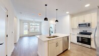 This well-designed single-floor residence offers two bedrooms, one bathroom, and a thoughtfully organized layout that maximizes functionality and comfort. The open-concept kitchen and living area serves as the heart of the home, featuring a large central island with a built-in sink, white quartz countertops, light wood cabinetry, a classic subway tile backsplash, and a full suite of stainless steel appliances. Striking black pendant lights over the island add a modern contrast to the bright, airy palette throughout.

The bedrooms are finished with warm wood-look luxury vinyl plank flooring, crisp white walls, and ceiling fans that enhance air circulation and comfort. Large windows invite an abundance of natural light while offering views of the surrounding landscaped exterior. The primary bedroom also features direct access to an outdoor area. Each bedroom is complemented by its own dedicated walk-in closet, outfitted with multiple clothing rods and shelving for ample storage.

The bathroom is elegantly appointed with light wood cabinetry, a dual-sink vanity with white countertops, matte black fixtures, a soaking tub, and a marble-tiled walk-in shower. A connecting hallway ties all rooms together efficiently. The exterior view reveals a contemporary multi-story building with lush tropical landscaping.
