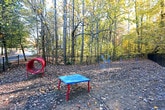 This outdoor dog park offers a dedicated, well-defined space for pets to roam and play in a natural setting. The area is fully enclosed by a sturdy black iron fence with a gated entry, providing a safe and secure environment for dogs of all sizes. The grounds feature a natural earth and leaf-covered surface that blends seamlessly with the surrounding woodland landscape, giving the space an organic, open-air feel.

Scattered throughout the park are blue agility and play structures, offering interactive opportunities for pets and their owners. A classic wooden-slatted bench with ornate iron legs is positioned along the fencing, providing a comfortable resting spot for visitors while they supervise their pets.

The park is framed by a dense canopy of mature deciduous and evergreen trees displaying rich autumn foliage in shades of gold, amber, and green, creating a scenic and tranquil atmosphere. A gently sloping hillside along one boundary adds natural topographic interest to the space. The surrounding residential buildings and parking areas are visible beyond the fence line, situating the park conveniently within a larger community. Overall, the dog park combines functional amenities with a naturally wooded ambiance, making it an inviting retreat for pets and their owners alike.