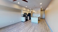 Located at 760 North Central Avenue in Moses Lake, Washington, this single-floor unit offers a thoughtfully designed open-concept layout with contemporary finishes throughout. The kitchen is a standout feature, boasting white cabinetry with sleek hardware, black appliances including a range, microwave, and refrigerator, and a large peninsula island topped with stone countertops that also houses a stainless steel sink. Pendant lighting and open floating shelves add a modern touch to the space. The kitchen flows seamlessly into a spacious living area with light wood-style flooring, recessed lighting, and a ceiling fan overhead — creating a bright, airy atmosphere. One bedroom features warm yellow accent walls, a large window with views of the surrounding neighborhood and balcony railing, and wood-style flooring, while a second bedroom is finished with patterned carpet and a ceiling fan. The bathroom is well-appointed with white cabinetry, a stone-topped vanity, a tub-shower combo with white tile surround, and wood-style flooring. Neutral tones, clean lines, and modern fixtures carry consistently throughout, giving the unit a cohesive and polished feel.