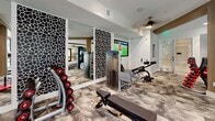 This single-level fitness center is designed to accommodate a comprehensive range of workout styles within a generously sized, open-plan layout. The space is divided into distinct training zones, each thoughtfully appointed with professional-grade equipment. A standout feature is the long, numbered turf sprint track that runs centrally through the facility, flanked by warm wood-wrapped structural columns that add an architectural accent to the otherwise neutral white interior.

The strength training area includes a multi-station cable machine, a full rack of color-coded dumbbells on curved storage stands, weight benches, and a dedicated functional training rig equipped with resistance bands, medicine balls, and a heavy bag. Large wall-mounted flat-screen TVs are positioned throughout, providing entertainment during workouts. Full-length mirrors paired with bold black-and-white decorative panels enhance the visual depth of the space.

The cardio zone features treadmills, an elliptical, and a stationary bike, positioned near large windows and glass partitions that allow natural light to filter in and offer views of adjacent areas. Patterned carpet throughout adds texture and a modern aesthetic. Ceiling fans and recessed lighting ensure consistent comfort and visibility across all zones.