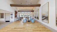 This generously proportioned single-floor space seamlessly blends a living room and dining area into one cohesive, open-plan environment. The architectural design is defined by soaring ceilings adorned with prominent exposed wood beams that add warmth and texture against crisp white walls. Light-toned wide-plank hardwood flooring runs throughout, enhancing the bright, airy atmosphere.

Natural light floods the interior through a series of clerestory windows positioned high along the walls, as well as multiple sets of black-framed French doors that provide outdoor access and views of the surrounding landscape. Elegant pendant lighting and recessed fixtures supplement the natural light beautifully.

The living area features multiple seating arrangements, including plush swivel chairs in soft blue and neutral tones grouped around low tables, offering a variety of intimate conversation areas. A standout textured accent wall in rich brown tones anchors one end of the room, complemented by a curated triptych of large-scale abstract artworks displayed in gold frames.

The dining zone includes a substantial white island-style counter with bar seating, backed by a floor-to-ceiling textured tile wall and integrated cabinetry — ideal for gatherings and casual dining. The overall ambiance is contemporary yet welcoming, with a refined coastal aesthetic achieved through a neutral palette, organic materials, and thoughtfully selected furnishings.