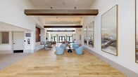 This generously proportioned single-floor space seamlessly blends a living room and dining area into one cohesive, open-plan environment. The architectural design is defined by soaring ceilings adorned with prominent exposed wood beams that add warmth and texture against crisp white walls. Light-toned wide-plank hardwood flooring runs throughout, enhancing the bright, airy atmosphere.

Natural light floods the interior through a series of clerestory windows positioned high along the walls, as well as multiple sets of black-framed French doors that provide outdoor access and views of the surrounding landscape. Elegant pendant lighting and recessed fixtures supplement the natural light beautifully.

The living area features multiple seating arrangements, including plush swivel chairs in soft blue and neutral tones grouped around low tables, offering a variety of intimate conversation areas. A standout textured accent wall in rich brown tones anchors one end of the room, complemented by a curated triptych of large-scale abstract artworks displayed in gold frames.

The dining zone includes a substantial white island-style counter with bar seating, backed by a floor-to-ceiling textured tile wall and integrated cabinetry — ideal for gatherings and casual dining. The overall ambiance is contemporary yet welcoming, with a refined coastal aesthetic achieved through a neutral palette, organic materials, and thoughtfully selected furnishings.