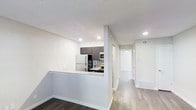 This single-floor space showcases a clean, modern design with a neutral white palette, light hardwood-style flooring, and recessed lighting throughout. The kitchen is a standout feature, equipped with dark espresso cabinetry, a mosaic tile backsplash, stainless steel appliances including a built-in microwave, and a peninsula topped with granite-style countertops. A full-size refrigerator anchors one side of the kitchen, providing ample storage and functionality. The peninsula serves as a natural divider between the kitchen and the adjoining living area, creating an open yet defined layout. A textured ceiling adds subtle dimension to the space. The hallway extending from the main area is bright and well-lit, with recessed lighting and white doors leading to additional rooms, suggesting a practical and organized floor plan. Trim details and clean baseboards throughout contribute to a well-finished aesthetic. The transition between the darker flooring near the kitchen and the lighter hardwood-style flooring in the hallway creates a subtle visual contrast, further defining the distinct zones within this efficiently designed single-floor space.