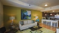 This single-floor space showcases a thoughtfully designed open-concept layout that connects the living area to the kitchen, creating a cohesive and functional environment. The living room is anchored by a bold olive-green accent wall adorned with a large-scale abstract painting in rich blues and greens, lending an artistic focal point to the space. A neutral-toned sofa with teal accent pillows sits atop a green botanical-patterned area rug, complementing the vibrant color palette throughout. A sleek, dark rectangular coffee table grounds the seating arrangement, while coordinating table lamps in blue add warm, ambient lighting. The ceiling features a distinctive curved architectural detail with a concrete finish, adding a modern industrial character to the room. Light wood-toned flooring extends throughout, providing warmth and continuity across the space. The adjacent kitchen is visible from the living area, offering dark cabinetry, pendant lighting, and bar-height seating along a counter, making it ideal for casual dining or entertaining. Overall, the space balances contemporary design elements with warm tones and curated décor to create an inviting, modern atmosphere.