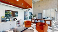 This single-floor commercial lobby and leasing space makes a strong architectural statement through a combination of industrial and contemporary design elements. The soaring double-height ceilings create an expansive, open atmosphere, punctuated by a bold orange-painted ceiling volume with exposed metallic ductwork that adds an industrial-chic character. The dramatic black-and-white checkered marble flooring anchors the space with a classic yet striking visual foundation.

Floor-to-ceiling windows along one wall flood the interior with abundant natural light while providing open views to the surrounding streetscape. A prominent brick accent wall rises to the full ceiling height, providing textural contrast against the clean white surfaces throughout.

The reception area features a substantial wooden front desk positioned centrally, alongside a secondary dark-wood workstation with ample counter space. A stylish lounge area incorporates tufted burnt-orange velvet barrel chairs paired with sculptural white side tables, creating an inviting atmosphere. Large-scale floral artwork adorns the walls, contributing to the vibrant aesthetic.

A well-equipped refreshment station with cabinetry, a beverage refrigerator, and a coffee setup is accessible near the entrance. Double glass doors lead directly to an adjacent garage or exterior area, enhancing connectivity and functionality throughout the space.