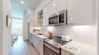 This thoughtfully designed single-floor residence combines modern finishes with functional living in a compact, well-organized layout. The kitchen is a standout feature, outfitted with stainless steel appliances including a flat-top electric range, over-the-range microwave, dishwasher, and refrigerator. Flat-panel cabinetry in a light wood tone is paired with high-gloss white upper cabinets and open shelving, creating a bright and contemporary feel. A white subway tile backsplash and light-toned countertops complete the polished look, all set atop warm hardwood-style flooring that runs throughout the space. The bedroom is inviting and filled with natural light, courtesy of large windows that bring in an abundance of daylight. White walls and clean lines keep the room feeling open and airy. The full bathroom features large-format grey tile surrounding the soaking tub and shower, complemented by a wood-toned vanity with a white countertop and a generous mirror. In-unit stacked laundry is also included, adding a practical convenience to the overall layout. Throughout the space, the consistent use of light tones, natural wood flooring, and modern fixtures creates a cohesive, contemporary ambiance that is both stylish and livable.
