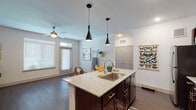 This single-story residence is designed with a modern, open-concept layout that seamlessly connects the kitchen and living areas. The kitchen serves as a central focal point, anchored by a large island with granite countertops and a stainless steel sink, illuminated by a pair of sleek black pendant lights. Dark espresso-toned cabinetry provides ample storage, while stainless steel appliances—including a refrigerator, range, and microwave—add a polished, contemporary finish. A herringbone tile backsplash adds a refined design detail along the main kitchen wall.

The open living area extends naturally from the kitchen, featuring dark hardwood-style flooring that runs throughout the unit, white walls, and a ceiling fan that promotes air circulation. Large windows with horizontal blinds allow generous natural light to fill the space, creating a bright and welcoming ambiance.

The bedroom is a quiet, well-proportioned room with a ceiling fan, a window for natural light, and the same consistent hardwood-style flooring. The bathroom features a granite-topped dark vanity with a large mirror, a tiled tub-shower combination, and matching flooring, maintaining the cohesive design aesthetic found throughout the home.
