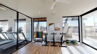 This single-floor yoga and fitness studio is designed with a clean, contemporary aesthetic that emphasizes openness, natural light, and functional versatility. The space is defined by its expansive floor-to-ceiling windows framed in sleek black metal, which flood the interior with abundant natural light and provide views of the surrounding outdoor terrace and skyline beyond.

The studio is organized into distinct functional zones. A dedicated cycling area features multiple stationary bikes arranged alongside a colorful medicine ball rack, with a ceiling fan overhead to maintain airflow during high-intensity sessions. Vaulted ceilings in this section enhance the sense of volume and airiness.

An adjacent strength training area includes a well-stocked kettlebell rack positioned beneath a large picture window, allowing natural light to illuminate the workout space. A mounted chalkboard display provides workout guidance nearby.

The open-floor yoga and group fitness zone is particularly spacious, finished with warm hardwood-style flooring that lends a welcoming, polished feel. Full-length mirrored walls visually expand the room, while a large flat-screen display mounted at one end supports guided workout programming. Stability balls and step platforms are conveniently accessible throughout.

A glass partition wall separates the yoga studio from an adjacent cardio room equipped with treadmills and additional fitness machines, maintaining visual connectivity while defining each area's purpose.