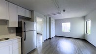 This single-floor space offers a clean, modern layout designed for comfortable everyday living. The main living area is anchored by sleek, dark hardwood-style flooring that runs throughout, complementing the neutral gray walls and contrasting black baseboards for a contemporary aesthetic. Exposed piping along the textured ceiling adds an subtle industrial character to the space.

The kitchen area is efficiently arranged, featuring white upper cabinetry and a full-size refrigerator, with direct sightlines into the living area, creating an open and connected feel. A window in the living area floods the space with natural light while framing views of the neighboring architecture, enhancing the sense of openness.

Multiple doorways lead to additional rooms, suggesting a practical and well-organized floor plan. Closet doors are conveniently situated near the living area, providing accessible built-in storage. The overall ambiance is bright, airy, and move-in ready, with a straightforward design that offers a neutral canvas adaptable to a variety of interior styles. The space strikes a balance between functional utility and modern minimalism, making it a versatile and inviting environment.