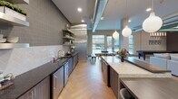 Located at 318 S Elkhart Ave, this single-floor community space for Stonewater at the Riverwalk showcases a thoughtfully designed interior that blends industrial and contemporary aesthetics. The welcoming entryway features striking herringbone-patterned flooring, a warm wood-slat ceiling, an interactive digital kiosk, and a built-in upholstered bench nook with textured geometric panels — setting an inviting tone from the moment you arrive.

The expansive open-concept main area is defined by soaring ceilings with exposed teal-accented ductwork and structural beams, creating an urban-loft atmosphere. Multiple seating arrangements — including a large sectional sofa, lounge chairs, and accent tables — provide versatile spaces for relaxation or casual gathering. Large floor-to-ceiling windows flood the area with natural light and offer views of the surrounding landscape.

The kitchen area is sleek and functional, featuring dark granite countertops, stainless steel appliances, a herringbone tile backsplash, floating metal shelves, and an adjacent island with bar seating beneath globe pendant lights. A dedicated conference room enclosed in glass offers a sophisticated setting with patterned wallcovering, a statement sputnik chandelier, a wall-mounted display screen, and seating for small group meetings.