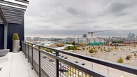The Sky Terrace at 651 W 42nd Ave in Denver, Colorado is a thoughtfully designed rooftop amenity space offering an elevated outdoor experience with panoramic views of the Denver skyline and surrounding urban landscape. The terrace is framed by a striking dark steel pergola structure with string lighting overhead, creating an inviting atmosphere for both daytime and evening use. Large-format light-toned tile flooring extends throughout the space, lending a clean, modern aesthetic.

A central lounge area anchors the terrace, featuring contemporary mint-upholstered seating arranged around a substantial concrete fire pit bowl — ideal for relaxed gatherings. The space transitions seamlessly into a covered section enclosed by corrugated dark metal panel walls and a translucent polycarbonate roof, providing shelter while maintaining an open, airy feel. Tall potted topiaries and ornamental greenery add natural accents throughout.

An outdoor dining area equipped with metal bistro-style tables and chairs is positioned to take full advantage of the expansive city views beyond the black metal mesh railing. A built-in grill station is also available, making the terrace well-suited for outdoor entertaining. Floor-to-ceiling glass sliding doors connect the covered area to the interior, ensuring easy access and excellent sightlines across the Denver metro area.