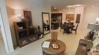 This single-floor residence showcases an open-concept design that integrates the living, dining, and kitchen areas into one cohesive, flowing space. The living area is anchored by a round, ornate coffee table atop neutral carpeting, complemented by a full-length floor mirror and dark wood shelving units that add both function and character. Recessed lighting and track lighting throughout ensure the space is well-illuminated, contributing to its warm and inviting ambiance.

The kitchen features dark cabinetry paired with stainless steel appliances, including a refrigerator and microwave, creating a sleek contrast against lighter walls and countertops. A raised breakfast bar provides casual seating, offering a natural transition point between the kitchen and the rest of the living space.

The dining area is furnished with a dark wood table and matching chairs, capable of accommodating several guests comfortably. A bold piece of artwork on the wall adds a striking focal point to this zone. The consistent use of dark wood tones across furniture pieces ties the entire space together, creating a cohesive, sophisticated aesthetic throughout.
