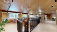 Located at 2080 FM 1102 in New Braunfels, Texas, this thoughtfully designed single-floor lounge space offers a refined and welcoming atmosphere rooted in natural materials and contemporary Texas Hill Country aesthetics. The interior is unified by light hardwood flooring that flows throughout, complemented by warm wood-paneled ceilings that lend a sense of intimacy and craftsmanship to the space.

The main lounge area is anchored by generously proportioned banquette seating upholstered in channeled fabric and leather, arranged alongside sculpted wood and marble-topped tables. A mix of lounge chairs and accent seating creates distinct conversational groupings, while layered area rugs define each zone within the open floor plan. Curated artwork, built-in shelving, and carefully placed greenery add depth and character throughout.

A standout architectural feature is the curved mezzanine overlook with sleek black metal railings and a wood-clad ceiling above, creating a dramatic upper vantage point. Pendant lighting descends from the ceiling, enhancing the space's ambient glow alongside wall sconces and track lighting. Large windows frame views of the surrounding Texas landscape, flooding the interior with natural light and connecting the space to its scenic outdoor surroundings. The overall ambiance is sophisticated yet approachable.