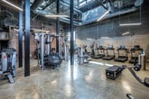 This fitness center in Memphis, Tennessee, delivers a compelling blend of industrial character and modern functionality. Housed within a repurposed building, the space retains its original exposed brick walls, weathered concrete floors, and visible ductwork overhead — all of which contribute to a raw, energetic atmosphere well-suited for an active facility.

The ceilings soar to impressive heights, creating an open, airy environment throughout the workout floor. Large multi-pane windows along one wall flood the space with abundant natural light, offering views of the surrounding cityscape while enhancing the overall ambiance. Suspended linear lighting fixtures provide consistent illumination across the entire floor.

The fitness floor is thoughtfully organized into distinct workout zones. A cardio section features a row of treadmills equipped with individual screens, alongside elliptical trainers and rowing machines. Strength training is well-represented with a power rack, a full dumbbell rack, weight plates, and a variety of cable and resistance machines. Functional training accessories include stability balls, medicine balls, resistance bands, and a plyo box. A flat-screen television is mounted on the wall for entertainment during workouts.

Additional amenities include a dedicated utility room and direct access via glass-panel entry doors. The combination of historic architectural detail and comprehensive modern equipment makes this fitness center a distinctive and capable training environment.