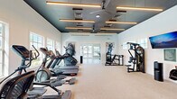 Located at 10196 SW Newberry Ave in Port Saint Lucie, Florida, this fitness center is a thoughtfully designed, single-floor facility divided into two distinct workout areas. The main exercise room features a striking teal-painted tray ceiling accented by linear pendant lighting and dual ceiling fans, creating a modern, energizing atmosphere. Soft, neutral-toned carpet lines the floor, and the space is outfitted with a comprehensive array of cardio equipment — including treadmills, ellipticals, and stair climbers — alongside a multi-functional cable strength machine and free weights. Wall-mounted televisions and large decorative artwork enhance the visual experience. A full-length mirror expands the sense of space and supports strength training. The secondary room offers a contrasting aesthetic with light hardwood flooring, warm neutral walls, and abundant natural light streaming through large grid-pane windows that frame views of a resort-style pool and outdoor amenity areas. This room features a Torque functional training unit with a heavy bag, kettlebells, and resistance attachments, as well as an indoor cycling station with a mounted display screen. French doors open to an outdoor terrace, further connecting the space to the Florida surroundings. Together, both rooms offer a comprehensive fitness experience suited for cardio, strength, and functional training.