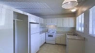 This single-floor space includes a kitchen designed for everyday functionality. The kitchen is outfitted with white cabinetry that lines the walls in an L-shaped configuration, providing ample storage through both upper and lower cabinets. Countertops in a neutral tone offer generous workspace, complementing the overall light color palette of the room. Essential appliances are included, among them a white electric range with an overhead exhaust hood, a full-sized refrigerator, and a sink positioned along one wall with a practical layout that supports ease of use. The flooring is a light-toned vinyl tile, contributing to the clean and low-maintenance character of the space. A textured ceiling adds subtle visual detail overhead, while a hanging light fixture provides overhead illumination. The neutral wall tones and white finishes throughout the kitchen create a bright, cohesive appearance. The layout is efficient, making good use of the available space and ensuring that the kitchen remains accessible and well-organized. Overall, the space presents a straightforward, functional design suited to everyday living.