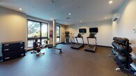 Located at 577 S Water St in Providence, Rhode Island, this well-appointed fitness center offers a comprehensive workout environment across a single, open-plan floor. The space is designed with a clean, modern aesthetic, featuring neutral-toned walls, recessed lighting, and durable rubber flooring throughout that provides both safety and comfort underfoot.

The main workout area is generously proportioned, with structural columns lending an architectural character to the open layout. Large, floor-to-ceiling windows line the perimeter, flooding the space with natural light and offering pleasant outdoor views of surrounding greenery. Wall-mounted mirrors in the free weights section enhance the sense of space and allow users to monitor their form.

The strength training zone is well-equipped with a full rack of dumbbells, weight benches, a cable machine, and a multi-functional power rack. A dedicated functional training area near the entrance houses a tiered rack of medicine balls and wall-mounted resistance bands, ideal for dynamic and core-focused workouts.

The cardio zone features multiple treadmills, a rowing machine, an air bike, and an elliptical, each paired with wall-mounted flat-screen TVs for entertainment during workouts. The entrance area is accessible via glass-paneled doors and includes a clean, open stretch zone. The overall ambiance is professional and inviting, making it a fully equipped facility suited for a wide range of fitness routines.