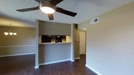 This single-floor residence offers a practical and well-organized layout designed for comfortable everyday living. The main living area features rich, dark hardwood flooring that extends throughout the space, providing a warm and grounded aesthetic. A ceiling fan with a built-in light fixture is centrally mounted on the textured ceiling, ensuring year-round airflow and ambient lighting.

The kitchen is defined by a raised pass-through counter that connects it to the living area, creating an open and interactive flow between the two spaces. Dark upper cabinetry lines the kitchen wall, offering ample storage, while a black appliance visible above the counter adds a cohesive, modern touch. A soffit above the kitchen area adds architectural definition to the space.

The walls are finished in a neutral warm beige tone, complementing the flooring and contributing to a calm, inviting atmosphere. A central hallway extends from the living area toward additional rooms, with a standard interior door visible at the end, suggesting private spaces beyond. HVAC ventilation is integrated into the ceiling, reflecting a fully climate-controlled environment. White baseboards throughout provide clean, finished transitions between the walls and flooring, adding a polished look to the overall interior.