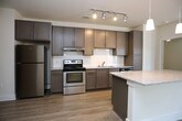 This well-designed single-floor residence offers a thoughtful layout that balances open communal spaces with comfortable private quarters. The heart of the home is an open-concept kitchen and living area, where light wood-toned flooring flows seamlessly underfoot. The kitchen is outfitted with gray shaker-style cabinetry, a classic white subway tile backsplash, stainless steel appliances including a refrigerator and range, and a granite-topped island with pendant lighting overhead. A dedicated pantry with built-in shelving adds generous storage. Track lighting provides functional illumination throughout the kitchen. The adjoining living area is bright and airy, featuring large windows that invite abundant natural light and views of surrounding greenery, complemented by a ceiling fan. The bedroom continues the warm, neutral palette with plush carpeting, a ceiling fan with light fixture, and a large window fitted with blinds. Storage is well addressed with a spacious walk-in closet equipped with wire shelving and hanging rods. Two bathrooms serve the residence, each featuring granite-topped dark vanities and tile flooring. The primary bathroom offers a wide vanity with a backlit mirror and direct access to the walk-in closet, while the secondary bathroom includes a tub-shower combination. Dark-stained interior doors throughout provide a refined contrast to the light walls and flooring.