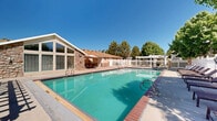 Located in Riverton, Utah, this outdoor community recreation area offers a well-appointed amenity space designed for relaxation and leisure. The centerpiece is a generously sized swimming pool with crystal-clear turquoise water, bordered by a wide concrete deck lined with lounge chairs — ideal for sunbathing beneath Utah's abundant blue skies. Adjacent to the main pool sits a circular hot tub with brick accents and easy-access handrails, providing a comfortable hydrotherapy option. A white pergola structure anchors one section of the deck, offering shaded seating and an outdoor dining area complete with a grill, perfect for social gatherings. The entire facility is enclosed by a classic white picket fence that defines the space while maintaining an open, airy feel. Mature trees surround the perimeter, providing natural shade and a lush, green backdrop throughout the warmer months. A single-story clubhouse building with striking floor-to-ceiling angled windows and stone facade accents overlooks the pool, blending architectural character with functional utility. The facility also includes a dedicated restroom and changing area. The overall ambiance is clean, welcoming, and thoughtfully maintained, making it an attractive community gathering space.