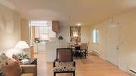 This single-floor space features an open-concept design that seamlessly integrates the living, dining, and kitchen areas into one cohesive environment. Light hardwood flooring runs throughout, lending warmth and continuity across the entire space.

The kitchen is positioned toward the rear and is equipped with white cabinetry, white appliances, and an overhead range hood. A peninsula-style counter separates the kitchen from the rest of the living area, offering both additional workspace and a natural visual boundary without disrupting the open flow.

Adjacent to the kitchen, the dining area accommodates a round table with upholstered chairs, creating an intimate setting for meals. A decorative feature wall with a mosaic-style tile accent adds a focal point and visual interest to this area, complementing the otherwise neutral color palette of white and warm beige walls.

The living area is positioned in the foreground and is furnished with accent chairs, contributing to a relaxed and functional layout. Natural light filters in through windows along the perimeter, brightening the interior. A baseboard heating unit runs along one wall, and the overall ambiance is clean, understated, and practical.