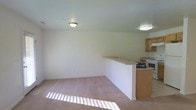 This single-floor residence offers a straightforward and functional layout designed for comfortable everyday living. The open-concept design seamlessly connects the living area to the kitchen, creating a cohesive and practical flow throughout the space.

The living area is finished with wall-to-wall carpeting in a neutral tone, providing a warm underfoot feel. Flush-mount ceiling lighting illuminates the room evenly, and natural light streams in through a window, casting bright patterns across the floor and adding a sense of openness to the space.

A half-wall partition defines the boundary between the living area and the kitchen, maintaining an open sightline while subtly separating the two zones. The kitchen features medium-toned wood cabinetry and is equipped with a range and microwave, offering essential cooking functionality. The cabinetry provides ample storage, keeping the space organized and efficient.

The walls throughout are finished in a clean, neutral palette that enhances the brightness of the interior and offers a versatile backdrop for a variety of furnishing styles. Overall, this space presents a clean, unpretentious layout that prioritizes practicality and ease of living.