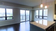 This single-floor residence showcases a thoughtfully designed open-concept layout that seamlessly connects the living and kitchen areas. Dark hardwood flooring runs throughout, providing a rich, contemporary contrast against the neutral-toned walls and ceiling.

The living area is bathed in natural light, courtesy of floor-to-ceiling sliding glass doors that lead to a private balcony with metal railing. The balcony offers elevated views of the surrounding cityscape and greenery, making it a standout feature of the space. An additional large window on the adjacent wall further enhances the bright, airy atmosphere while framing additional urban views.

The kitchen area is defined by a substantial granite-topped island or peninsula, featuring a speckled stone surface with warm golden tones that adds both functionality and visual interest. A modern pendant light hangs above, providing focused task lighting. The open sightlines from the kitchen to the living space ensure a connected, social layout ideal for everyday living and entertaining. Overall, the unit balances modern finishes with a clean, functional design.