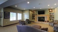 This single-floor communal lounge offers a comfortable and inviting atmosphere designed for relaxation and gathering. The space is anchored by a striking stone-surround fireplace, which serves as the focal point of the room and adds a warm, classic character to the interior. Mounted above the fireplace is a flat-screen television, making the area well-suited for both quiet evenings and social viewing. Adjacent to the fireplace, a built-in wood-toned bookshelf provides storage and display space, adding both function and a homey aesthetic to the room. The seating arrangement includes a mix of upholstered armchairs and sofas in muted tones, thoughtfully arranged to encourage conversation and comfort. Recessed lighting in the textured ceiling provides even, ambient illumination throughout the space. Natural light filters in through multiple windows along one wall, brightening the neutral green and beige color palette. The carpeted flooring contributes to the overall warmth and acoustic comfort of the room. A doorway visible along one wall suggests easy access to additional areas. Overall, this lounge balances a relaxed residential feel with practical amenities, creating a welcoming environment for everyday use.