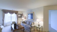 This single-floor residence presents a thoughtfully designed living room that combines traditional elegance with comfortable functionality. The living room is centered around a generously sized blue-striped sofa adorned with accent pillows, complemented by a matching armchair that creates an inviting seating arrangement. A richly carved wooden coffee table serves as the focal point of the seating area, while a glass-topped side table with a metal frame adds a refined accent on the opposite end.

The room benefits from natural light streaming through a large window dressed with layered drapes featuring decorative valances in warm gold tones, adding a classic, formal touch to the space. The walls are finished in a soft, neutral palette that enhances the airy ambiance. A gold-framed artwork prominently displayed on the main wall adds visual interest and a sense of sophistication.

Tasteful table lamps on either side of the room provide warm ambient lighting, while framed artwork arranged along one wall contributes to the curated, traditional aesthetic. The smooth, light-colored ceiling and neutral flooring tie the space together cohesively, creating a welcoming and polished atmosphere throughout.
