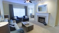 This single-floor residence showcases a well-appointed, open-concept layout that seamlessly connects the living and dining areas. The living room serves as the focal point of the space, centered around a tile-surround fireplace with a white mantel and a built-in recessed mirror above, adding depth and elegance to the room. Crown molding runs along the ceiling, lending a refined architectural detail throughout. A ceiling fan with a decorative light fixture provides both function and style overhead, complemented by a track lighting system that allows for flexible illumination.

Large windows dressed with layered blinds and patterned draping flood the space with natural light, creating a bright and welcoming atmosphere. The neutral-toned carpeting extends throughout, providing a warm, cohesive foundation. The living area comfortably accommodates a sofa, a pair of navy accent chairs, and a rustic wood coffee table, offering a balanced blend of comfort and style. Just beyond, the dining area features a table and chairs, making the transition between living and dining spaces fluid and functional. The overall ambiance is calm, polished, and inviting.