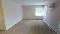 This single-floor space offers a clean, functional layout designed with simplicity and comfort in mind. The interior is finished with neutral beige walls and wall-to-wall carpeting in a warm, sandy tone, creating a cohesive and understated aesthetic throughout. The living area is generously proportioned, providing ample room for a variety of furniture arrangements. A large window fitted with white horizontal blinds allows abundant natural light to fill the space, while framing a pleasant view of surrounding greenery — a welcome touch of nature from within. Baseboard heating units are installed along the exterior walls, ensuring efficient and consistent warmth during colder months. The overall design is straightforward and practical, with smooth ceilings and clean wall surfaces that offer a versatile blank canvas for personalization. The neutral palette and carpeted floors contribute to a quiet, comfortable ambiance well-suited for everyday living. This space presents a solid foundation for those seeking a move-in ready environment with classic, low-maintenance finishes.