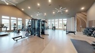 Located at 6615 Cerris Dr in Charlotte, NC, this fitness center offers a generously sized, single-floor exercise space designed to support a full range of workout routines. The room features soaring vaulted ceilings that lend an open, airy atmosphere, complemented by large windows along multiple walls that flood the interior with natural light and provide views of the surrounding community and pool area. Oversized ceiling fans ensure comfortable airflow throughout the space.

The flooring consists of light-toned wood-style panels that give the gym a clean, modern aesthetic. The cardio section is well-appointed with multiple treadmills and elliptical machines, each positioned to take advantage of the expansive window views. A cable and multi-functional strength training station anchors the center of the floor, while a dedicated free weights area features a full rack of dumbbells, a power rack with barbells, and a wall-length mirror for form monitoring. Additional selectorized weight machines provide targeted muscle group training options.

A standout artistic mural with flowing neutral tones adorns one accent wall, adding a dynamic visual element to the space. Built-in shelving along one wall provides organized storage, and a wall-mounted television enhances the overall workout experience. The facility is accessed through double glass doors, reinforcing the bright, welcoming character of this thoughtfully designed fitness space.