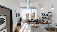 This single-floor loft-style residence showcases a compelling blend of industrial character and contemporary design throughout its open layout. Soaring ceilings with exposed silver ductwork establish an authentic converted-building aesthetic, while large, black-framed factory-style windows flood every room with abundant natural light and frame urban rooftop views.

The living area is generously proportioned, anchored by rich hardwood-style flooring and designed for comfortable gathering. Sheer white curtains soften the industrial windows, complementing the open, airy atmosphere. An open-concept kitchen sits adjacent to the living space, featuring granite countertops, white cabinetry with subway tile backsplash, stainless steel appliances, and ample preparation space. A granite-topped island or peninsula provides a natural transition between the kitchen and living areas.

The bedroom benefits from the same oversized factory windows, allowing natural light to pour in alongside views of surrounding brick architecture. The bathroom features a clean, bright tub-shower combination with a minimalist finish.

Striking matte black interior doors with frosted glass panels add a bold design contrast to the white walls throughout, while the exposed mechanical elements overhead reinforce the loft's distinctive industrial-modern identity.